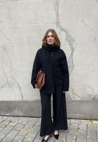 Vrouw met golvend haar, gekleed in een zwarte jas, brede broek en sjaal, die een bruine handtas vasthoudt, staande op een met bladeren bedekte stoep bij een gekraakte stenen muur.