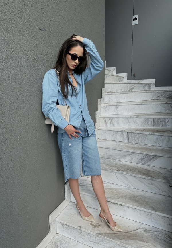 Camicia in denim azzurro chiaro con colletto, abbinata a pantaloni shorts in denim azzurro chiaro fino al ginocchio. Indossa scarpe beige a punta e tiene una borsa color crema.