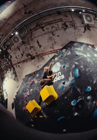 Persona che scala una parete di bouldering indoor con prese colorate e due grandi appoggi per i piedi a forma di cubo giallo.
