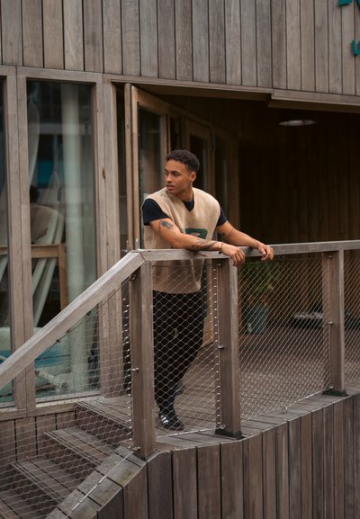 Chaleco de punto beige sobre una camiseta negra, de pie en una terraza de madera con barandilla. El fondo presenta grandes puertas de cristal y paredes de madera.