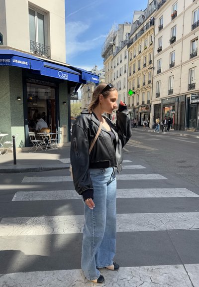 Una chaqueta bomber de cuero negro con puños acanalados, combinada con vaqueros de pierna ancha de color azul claro, destaca en el fondo de una calle parisina.
