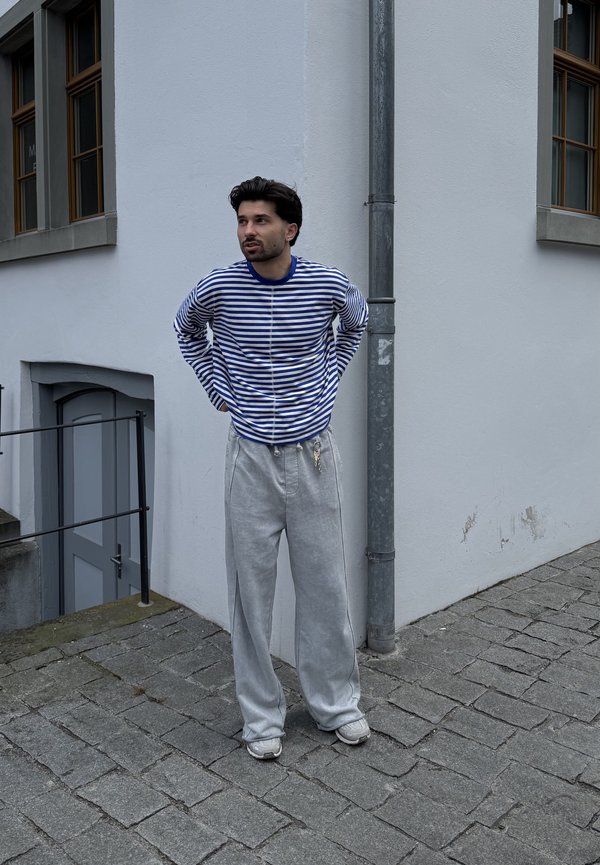 Homme en chemise à manches longues rayée bleu et blanc et pantalon gris, debout à un coin de rue pavée près d'un bâtiment blanc avec des fenêtres.