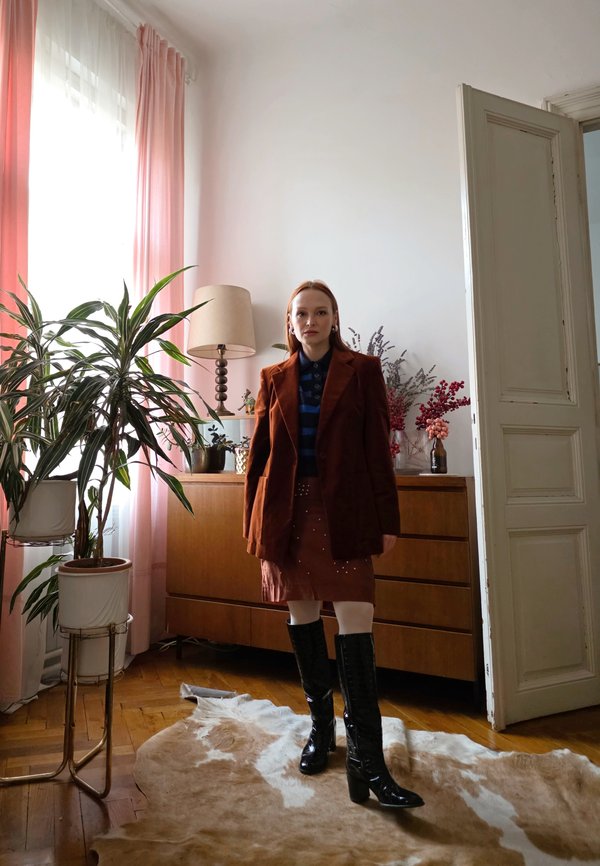 Femme en blazer marron, jupe et bottes noires, debout sur un tapis en peau de vache dans une pièce lumineuse avec des plantes, une commode en bois et une porte ouverte.