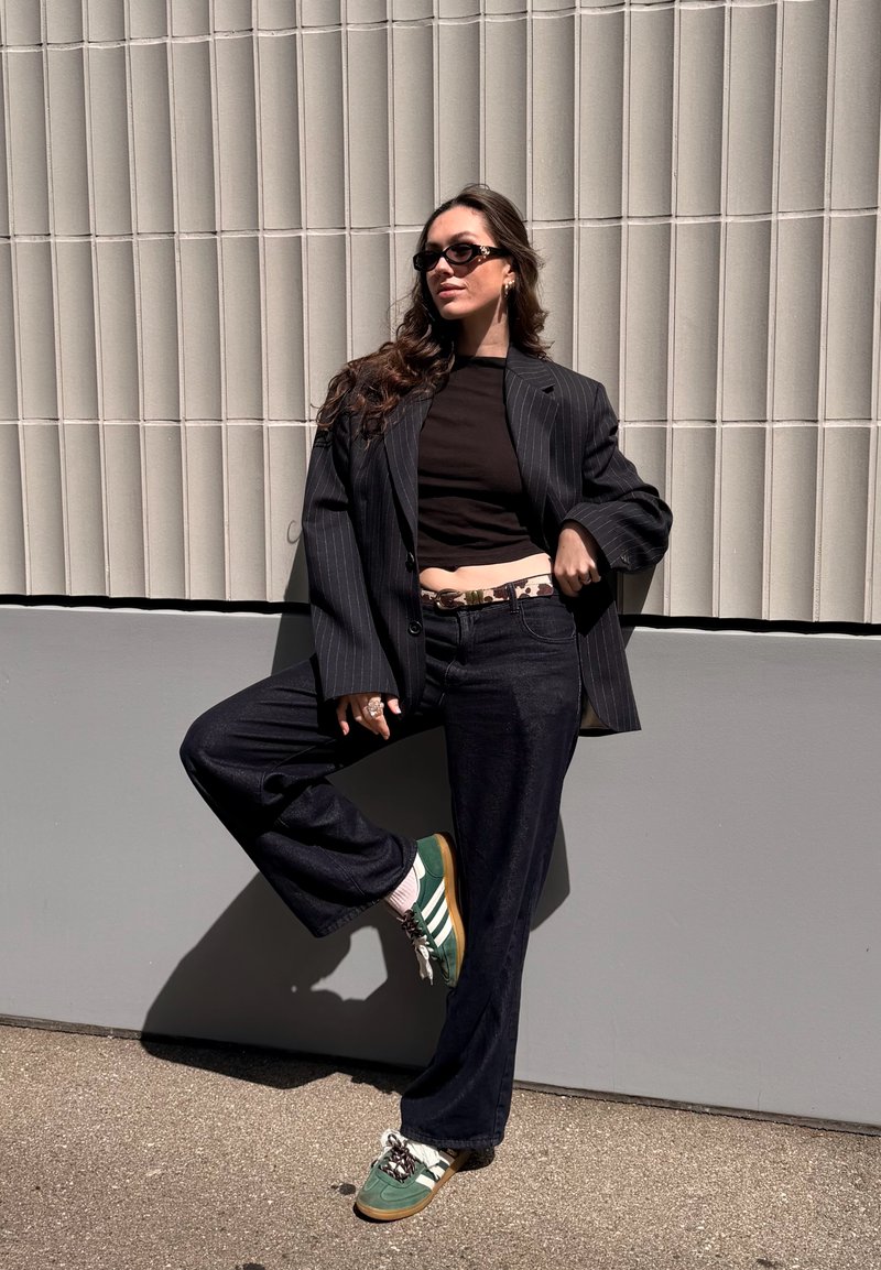 Femme portant des lunettes de soleil, un blazer noir à fines rayures, un haut noir, un jean foncé et des baskets vertes, posant devant un mur texturé en extérieur.