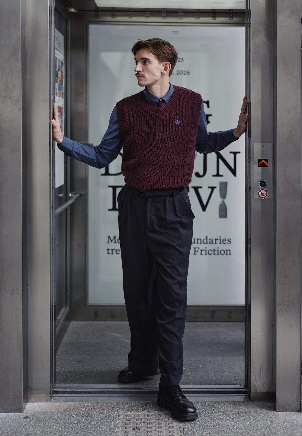 Gilet bordeaux en tricot porté sur une chemise bleu foncé, avec un pantalon noir plissé et des chaussures noires. L'homme se tient dans un ascenseur moderne.