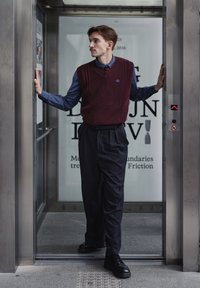 Gilet bordeaux en tricot porté sur une chemise bleu foncé, avec un pantalon noir plissé et des chaussures noires. L'homme se tient dans un ascenseur moderne.