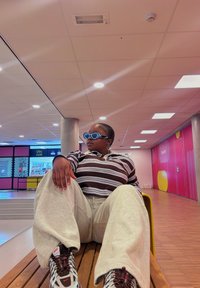 Camicia a maniche lunghe a righe marroni e bianche, pantaloni della tuta grigi chiari, occhiali da sole blu oversize e sneakers a fantasia. Sfondo con panchina in legno.