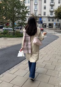Un cappotto lungo beige, una sciarpa a scacchi in toni tenui appoggiata sulla spalla, una borsa a mano rosa chiaro, jeans a zampa d'elefante blu, che tiene in mano una busta di carta bianca.