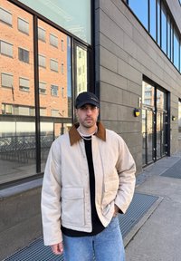 Beige jakke med brun cordfløyelskrage, to frontlommer, båret over en svart genser. Lyseblå jeans er synlige, står utendørs.