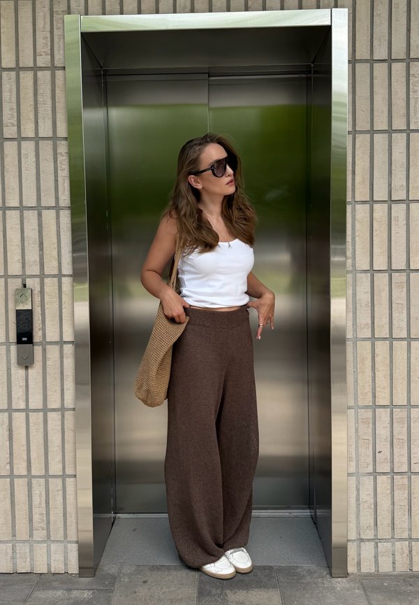 Donna con occhiali da sole, top bianco e pantaloni marroni in piedi in un ascensore aperto in acciaio inox con una borsa intrecciata sulla spalla.