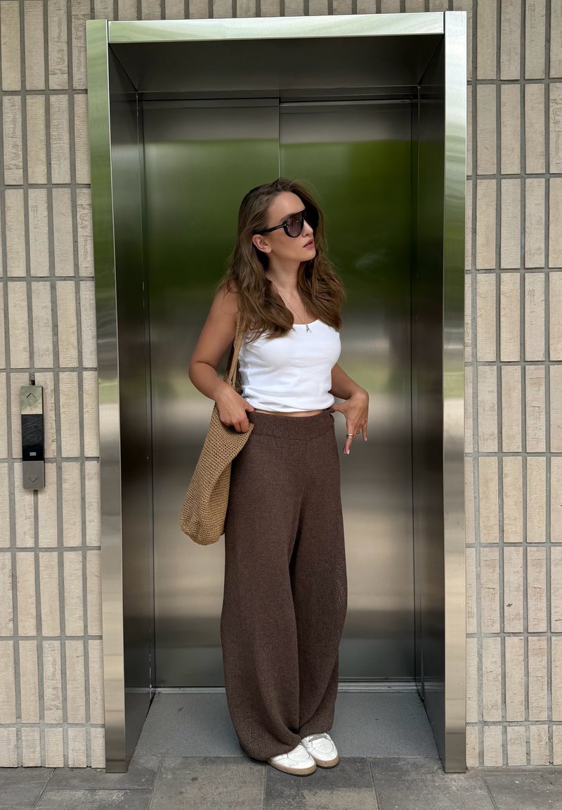 Femme portant des lunettes de soleil, un haut blanc et un pantalon marron, debout dans un ascenseur ouvert en acier inoxydable avec un sac tressé sur l'épaule.