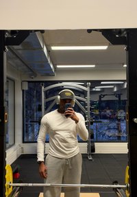 Homme portant une chemise blanche à manches longues, un pantalon de survêtement gris, une casquette noire et des écouteurs, prenant un selfie dans un miroir dans une salle de sport avec du matériel de musculation.