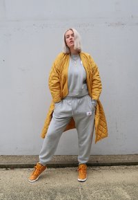 Gluosnė geltona ilga striukė, dėvima ant pilkos sportinės palaidinės ir atitinkamų sportinių kelnių. Oranžiniai sportbačiai su baltais akcentais. Paprasta fono.