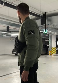 Grüne Puffjacke mit geripptem Kragen und Patch-Logo auf dem linken Ärmel. Schwarze Hüfttasche, die quer über den Körper getragen wird. Innenaufnahmesetting in einer Parkgarage.