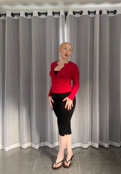 Mujer con cabello rubio que lleva un cárdigan rojo, leggings negros recortados y sandalias de dedo, de pie sobre un suelo de baldosas frente a unas cortinas grises.