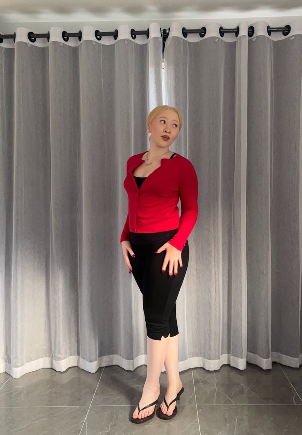 Kvinde med blondt hår iført rød cardigan, sorte cropped leggings og klipklappere står på flisebelagt gulv foran grå gardiner.