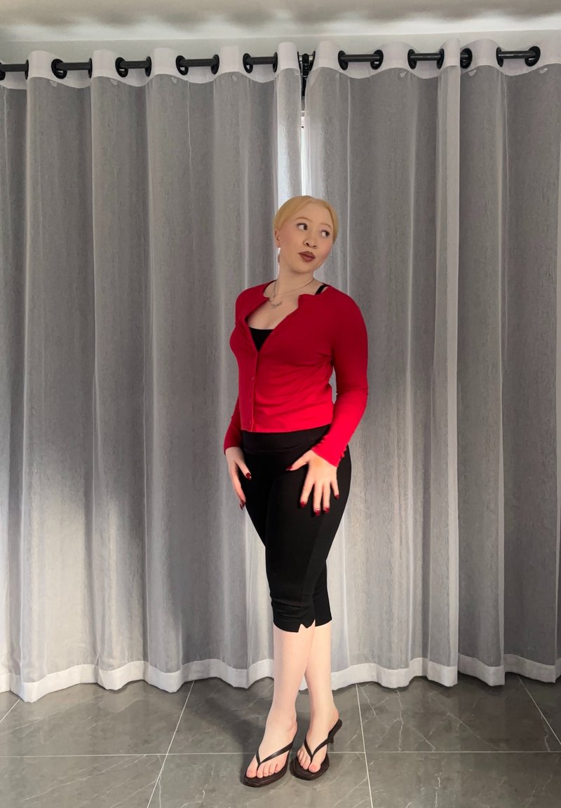 Femeie cu păr blond, purtând un cardigan roșu, leggings scurți negri și șlapi, stând pe un podea cu gresie în fața unor perdele gri.