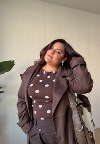 Brauner Wildleder-Trenchcoat über einem braunen gepunkteten Oberteil, kombiniert mit braunen Hosen. Eine Tasche mit fellartiger Textur und silberner Hardware wird getragen.