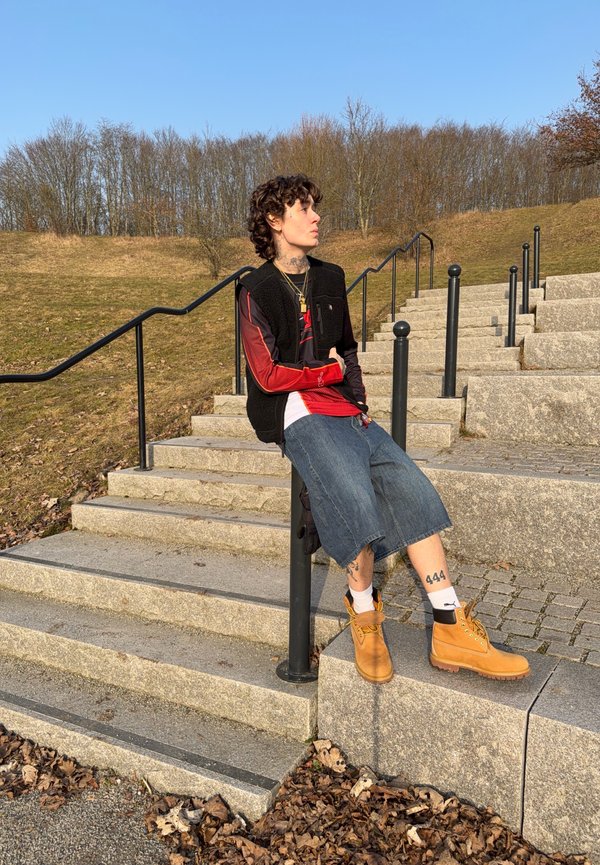 Osoba s kučeravými vlasmi, oblečená v čiernej veste, červenej dlhorukávovej košeli, rifľových kraťasoch a svetlohnedých topánkach, sediaca na stĺpe pri kamenných schodoch vonku.