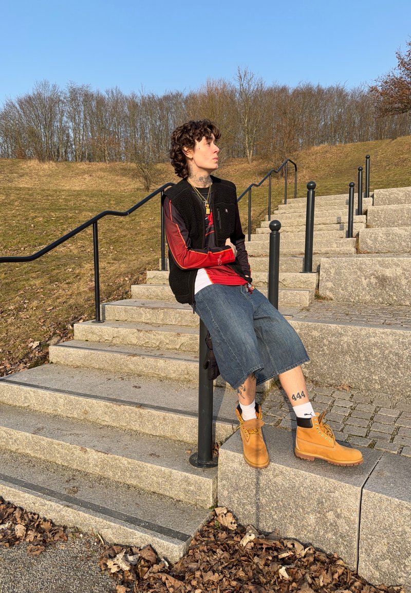 Osoba s kovrčavom kosom nosi crni prsluk, crvenu majicu s dugim rukavima, traper kratke hlače i smeđe čizme, sjedi na stupu pored kamenih stepenica na otvorenom.