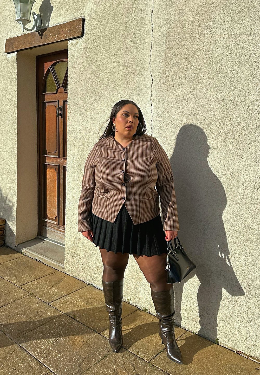 Femme portant une veste marron à carreaux, une jupe plissée noire et des bottes montantes, tenant un sac à main noir, debout contre un mur beige près d'une porte en bois.