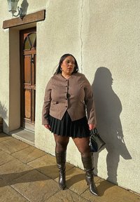 Femme portant une veste marron à carreaux, une jupe plissée noire et des bottes montantes, tenant un sac à main noir, debout contre un mur beige près d'une porte en bois.