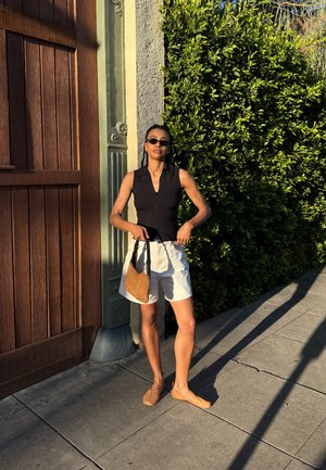 Mujer con gafas de sol, top negro sin mangas, pantalones cortos blancos y zapatos marrones, está de pie en la acera sosteniendo un bolso marrón junto a una puerta de madera y un seto verde.