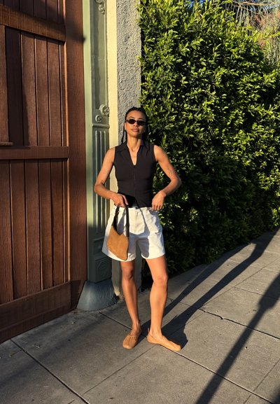 Mujer con gafas de sol, top negro sin mangas, pantalones cortos blancos y zapatos marrones, está de pie en la acera sosteniendo un bolso marrón junto a una puerta de madera y un seto verde.