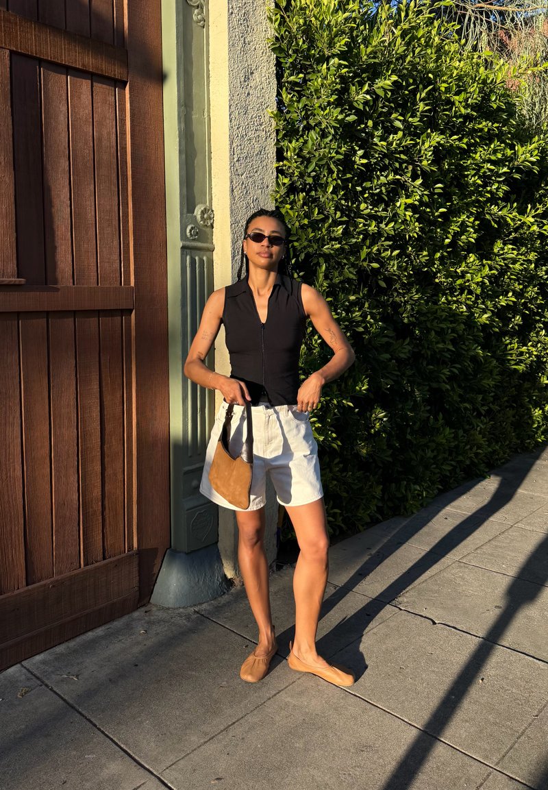 Kobieta w okularach przeciwsłonecznych, czarnej bluzce bez rękawów, białych szortach i brązowych butach stoi na chodniku, trzymając brązową torebkę obok drewnianych drzwi i zielonego żywopłotu.