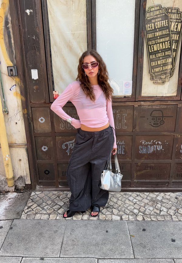 Femme aux cheveux ondulés portant un haut rose à manches longues, un pantalon noir oversize et des lunettes de soleil roses, debout dans une rue pavée tenant un sac à main argenté.