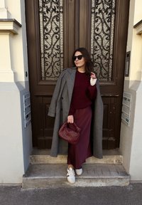 Manteau gris, pull bordeaux, longue jupe bordeaux, chaussures blanches avec accents rouges, sac à main tissé bordeaux, debout près d'une porte en bois ornée.
