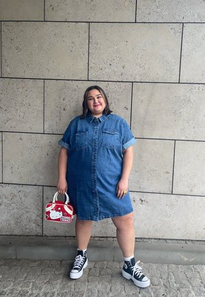 Vestido de camisa de denim con mangas cortas dobladas, bolsillos frontales y un lavado claro. Combinado con zapatillas negras de caña alta y un bolso de Hello Kitty.