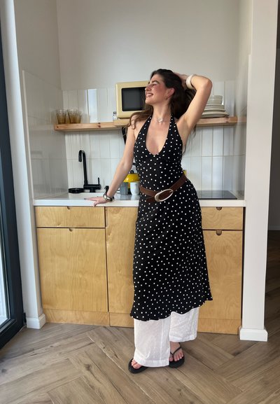 Vestido halter de lunares negros, pantalones blancos con adornos, cinturón marrón con hebilla circular, ambientado en una cocina moderna con muebles de madera.