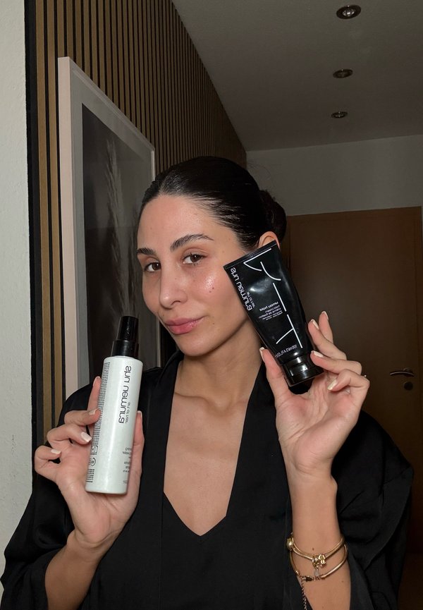 Bottiglia spray bianca con tappo nero, con etichetta "Amruta", in una mano e tubo di crema nero con tappo d'argento nell'altra, entrambi tenuti contro uno sfondo neutro.