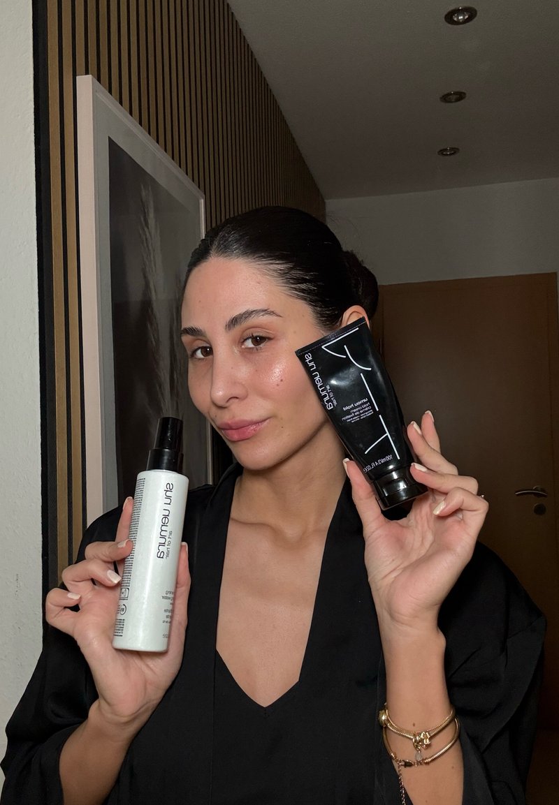 Bouteille pulvérisateur blanche avec un couvercle noir, étiquetée "Amruta", dans une main, et un tube de crème noire avec un bouchon argenté dans l'autre, tous deux tenus devant un fond neutre.