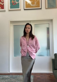 Camicia rosa oversize con bottoni e maniche lunghe, abbinata a una gonna satinata slim di colore neutro. Texture liscia e vestibilità rilassata.