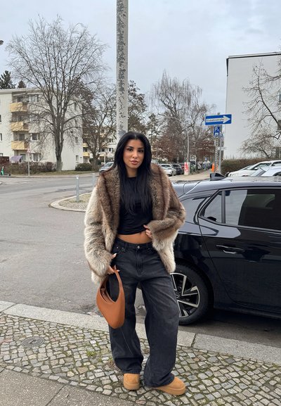 Abrigo de piel sintética en tonos marrones, top negro, jeans negros holgados, bolso marrón y botas color arena. Entorno urbano con árboles y coches al fondo.