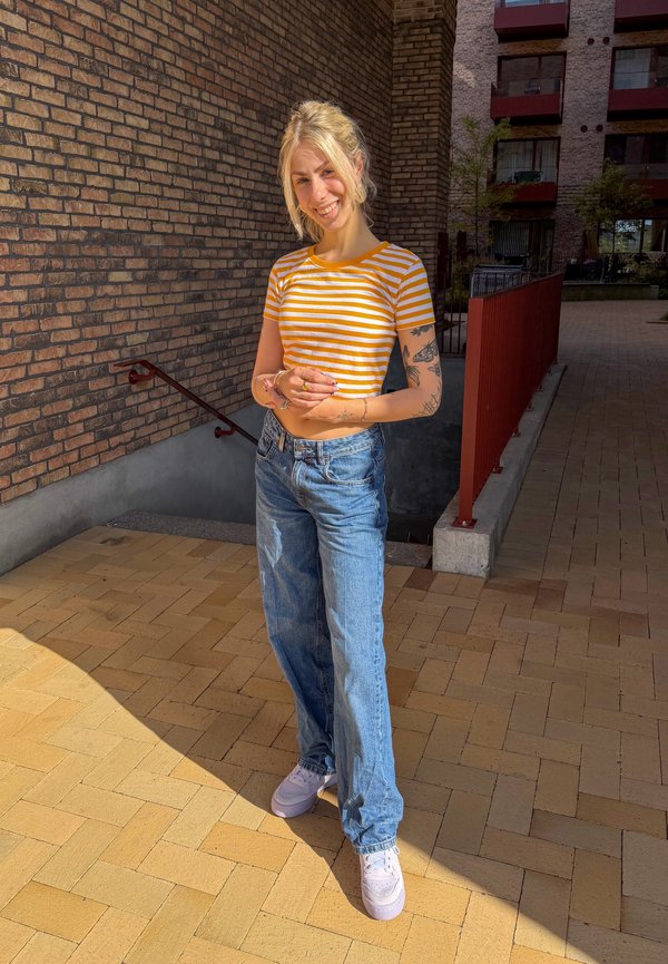 Mujer joven con cabello rubio, camiseta a rayas naranja y blanca, vaqueros azules y zapatillas blancas, de pie afuera cerca de una pared de ladrillo y escaleras.