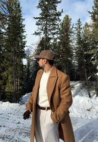 Άνδρας φοράει καφέ παλτό, καπέλο από τουίντ και γυαλιά ηλίου, στέκεται σε χιονισμένο δάσος με αειθαλή δέντρα και βουνά στο φόντο.