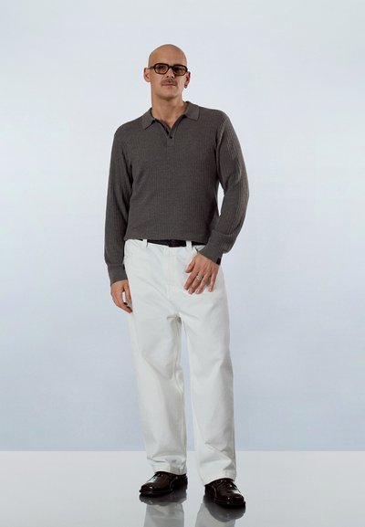 Hombre calvo con gafas, vistiendo un polo de manga larga gris, pantalones blancos, cinturón negro, anillos y zapatos marrón oscuro, de pie sobre una superficie reflectante.