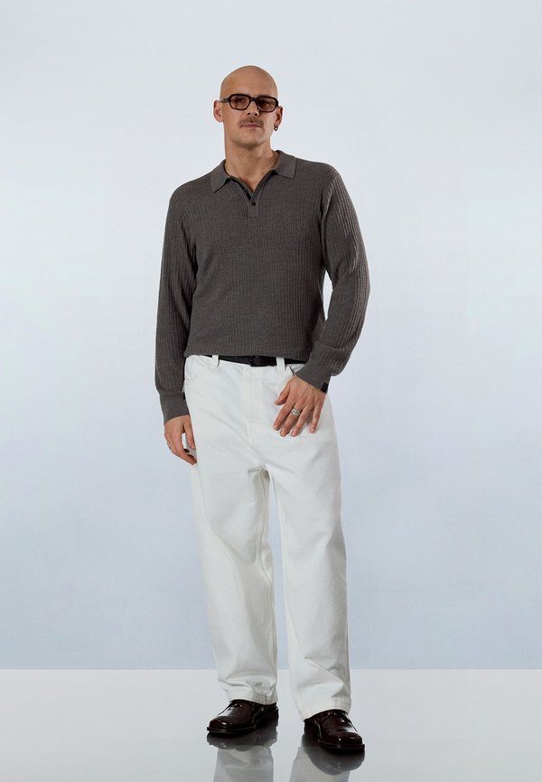 Plešatý muž s okuliarmi, oblečený v sivom polokoši s dlhým rukávom, bielych nohaviciach, čiernom opasku, prsteňoch a tmavohnedých topánkach, stojaci na lesklej ploche.