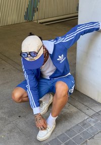 Tuta Adidas blu con strisce bianche, abbinata a sneaker bianche. L'individuo ha tatuaggi e indossa un cappellino chiaro e occhiali da sole.
