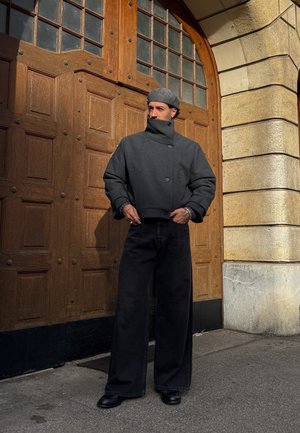 Hombre con chaqueta gris de cuello alto, boina gris, pantalones anchos negros y zapatos negros, de pie frente a una gran puerta de madera y pared de piedra.