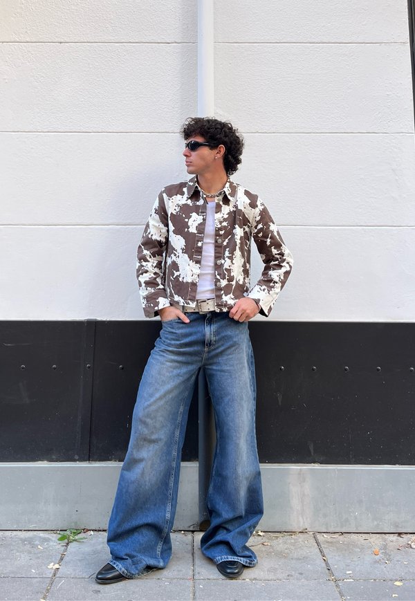 Casaco de padrão castanho e branco, camisa branca, calças de ganga azuis de perna larga, sapatos pretos e óculos de sol, em pé contra uma parede neutra.