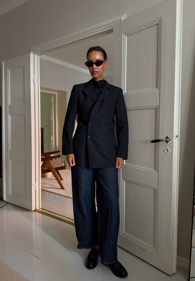 Blazer negro de doble botonadura con mangas largas, combinado con vaqueros azules holgados y zapatos negros. Los accesorios incluyen unas pequeñas gafas de sol negras.