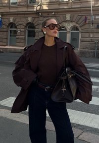 Brun oversized trenchcoat, mørkebrun genser, høyt liv mørke jeans, svart belte, stor håndveske, og oversized solbriller.