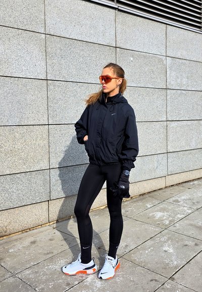 Chaqueta deportiva negra con capucha, mallas negras y zapatillas blancas con detalles en naranja. Lleva gafas de sol y guantes negros. Escenario urbano.