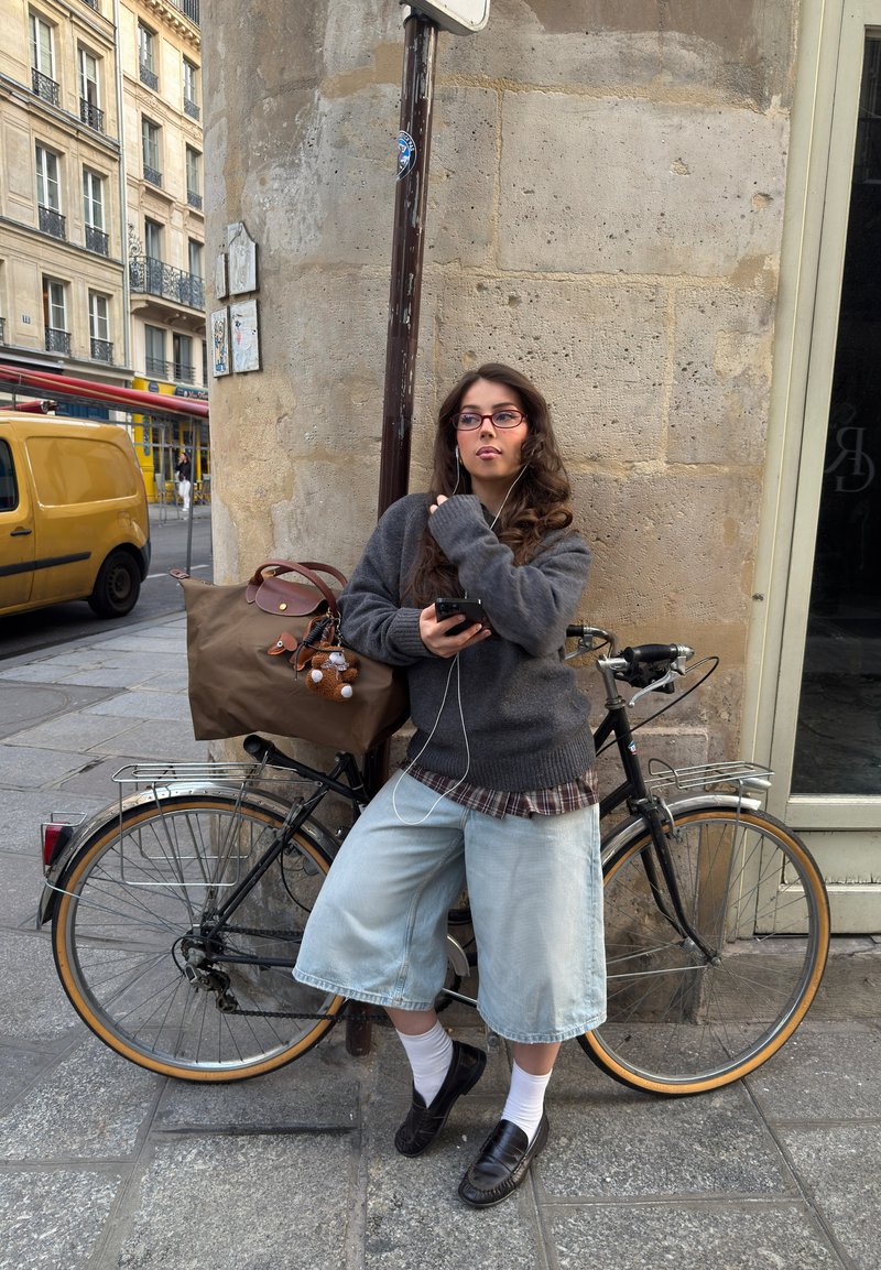 Junge Frau mit Brille und Kopfhörern lehnt an einem Fahrrad, hält ein Handy und steht auf einem Bürgersteig in der Stadt nahe einem gelben Van.