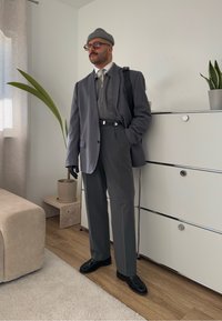 Harmaa räätälöity puku, jonka alla on vetoketjullinen villapaita, mustat käsineet ja neulottu pipo. Seisoo kasvin ja huonekalun vieressä minimalistisessa ympäristössä.