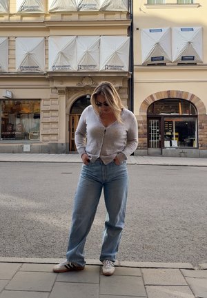 Cárdigan gris claro con botones, combinada con vaqueros de tiro alto en azul claro. Zapatillas con rayas en blanco y negro. Fondo urbano.